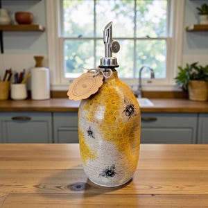 May include: A ceramic oil dispenser with a speckled beige base and a yellow honeycomb pattern featuring black bee illustrations. The bottle has a silver metal pour spout and a scalloped brown paper tag tied with twine around the neck.