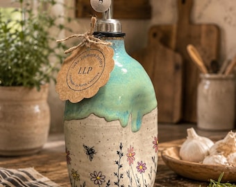 Puede incluir: Un dispensador de aceite de cerámica con un esmalte turquesa que gotea en la parte superior y una base crema moteada con tallos negros pintados a mano, flores rosas y amarillas, y una abeja. Una etiqueta de papel marrón dice Lake Lismore Pottery LLP.
