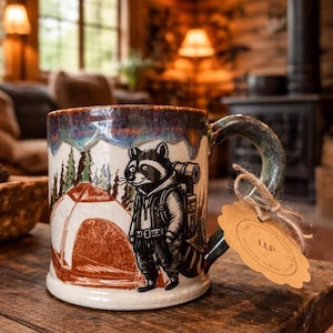 May include: A ceramic mug featuring a raccoon illustration with a backpack, set against a backdrop of a tent and trees. The mug has a brown, blue, and green glazed rim and handle. A tag is attached.