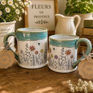 Puede incluir: Dos tazas de cerámica con diseños florales. Las tazas tienen una base blanca con flores coloridas y un borde y asa turquesa. Cada taza tiene una etiqueta atada al asa. Las tazas están sobre una superficie de madera.