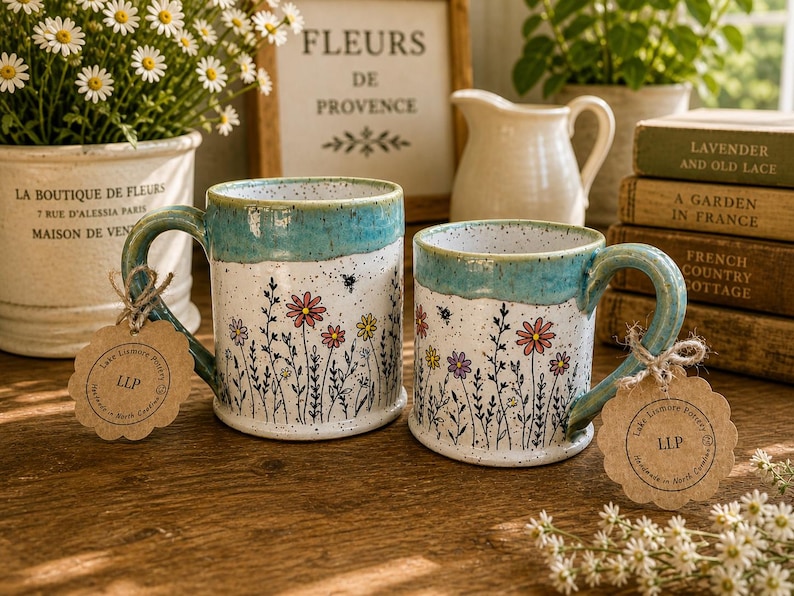 May include: Two ceramic mugs with floral patterns. The mugs feature a white base adorned with colourful flowers and a turquoise rim and handle. Each mug has a tag attached to the handle. The mugs are on a wooden surface.