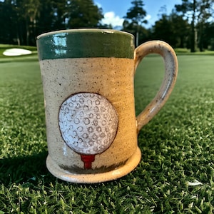May include: A handmade ceramic mug with a golf ball design. The mug features a tan body with a green rim and handle. A white golf ball on a red tee is painted on the front. The mug is sitting on green grass.