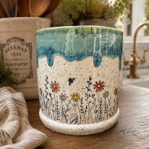 May include: A cylindrical ceramic utensil holder with a speckled cream base featuring hand-painted black line art of wildflowers and a small bee. The top rim has a thick, glossy blue-green glaze that drips down over the speckled surface.