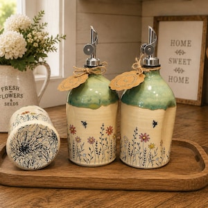 Puede incluir: Dos dispensadores de aceite de cerámica con vertedores plateados y etiquetas de papel marrón, base crema con flores silvestres pintadas a mano, abejas y esmalte verde. Un tarro de cerámica a juego con un boceto floral negro sobre bandeja.