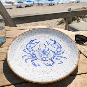 Puede incluir: Un plato redondo de cerámica con un diseño de cangrejo azul. El plato tiene una superficie blanca moteada y un borde marrón. Está sobre una mesa de madera, con una escena de playa al fondo.