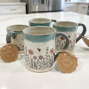 May include: Four handmade ceramic mugs with a speckled white base, teal rim and handle, and floral design. Each mug has a tag tied to the handle. The mugs are decorated with colorful flowers and bees.