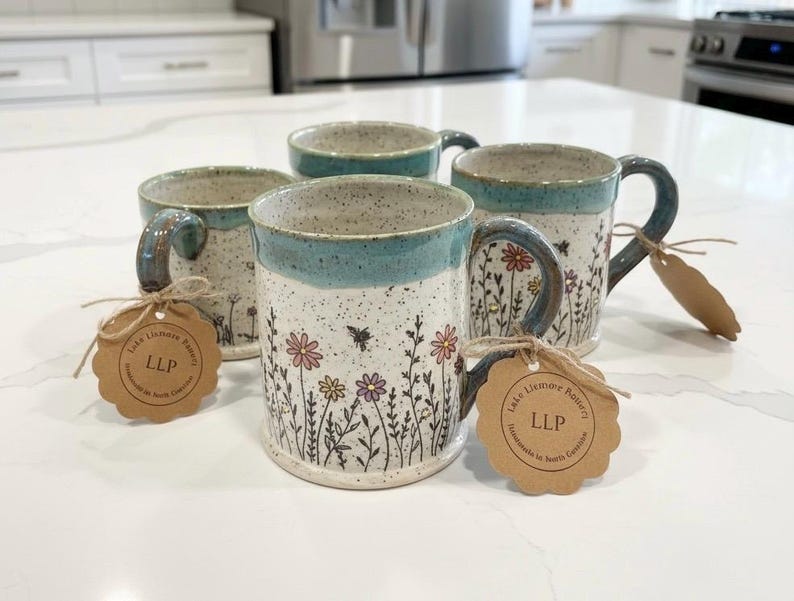 May include: Four handmade ceramic mugs with a speckled white base, teal rim and handle, and floral design. Each mug has a tag tied to the handle. The mugs are decorated with colorful flowers and bees.