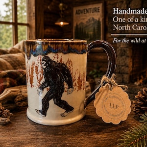 May include: A ceramic mug featuring a black Bigfoot silhouette and orange pine trees against a cream background, with a dark blue and brown drip glaze rim. A scalloped brown paper tag reads Lake Lismore Pottery, LLP, Handmade in North Carolina.