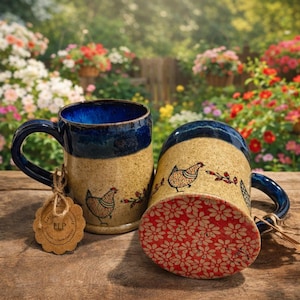 Puede incluir: Dos tazas de cerámica con asas y bordes azules, con una base beige con una ilustración de gallina y un estampado floral rojo en la parte inferior. Una taza está en posición vertical, la otra está de lado. Una etiqueta cuelga de una taza.