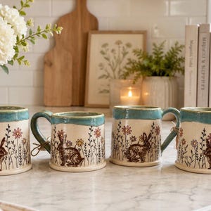 May include: Four ceramic mugs with a cream base, featuring a brown rabbit and floral design. Each mug has a teal rim and handle, tied with twine. The mugs are part of a spring-themed collection.