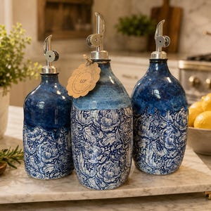 Puede incluir: Tres dispensadores de aceite de cerámica con un patrón floral azul y blanco. Cada botella presenta una parte superior esmaltada en azul oscuro liso y una base blanca con intrincados dibujos de flores en azul. Un vertedor de metal plateado está fijado a cada una.