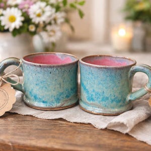 May include: Two ceramic mugs with a speckled turquoise and blue exterior glaze and a vibrant pink interior. Each mug features a sturdy handle and a scalloped brown paper tag tied with twine, displaying the text LLP and other printed details.