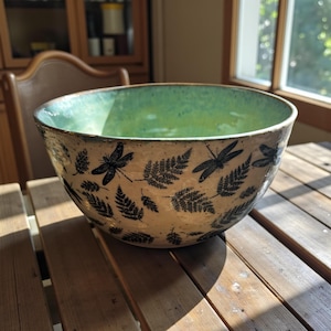 May include: A large ceramic bowl with a light beige exterior decorated with black dragonfly and fern designs. The interior is a gradient of green hues. The bowl sits on a wooden table, showcasing its handcrafted nature.