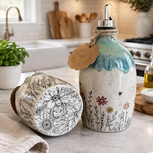 Op de afbeelding: Twee keramische oliedispensers met een crèmekleurige basis, handgetekende zwarte bloemmotieven en kleine kleurrijke bloemen. De rechtopstaande dispenser heeft een blauw geglazuurde bovenkant, een metalen schenktuit en een bruin papieren label.