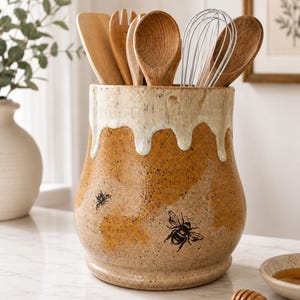 Könnte beinhalten: Ein Keramik-Utensilienhalter mit Honig-Thema. Der Halter ist beige mit einem Bienenwabenmuster und zwei schwarzen Bienenillustrationen. Er hat eine weiße, tropfende Glasur am oberen Rand und enthält Holzlöffel, eine Gabel und einen Schneebesen.