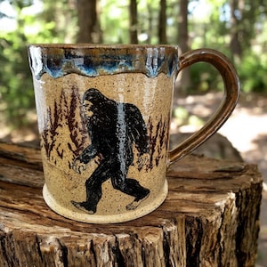 Puede incluir: Una taza de cerámica con un asa marrón y un cuerpo beige. La taza presenta una ilustración negra de Bigfoot caminando por un bosque. El borde tiene un esmalte azul y marrón.