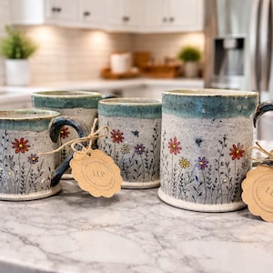 Może przedstawiać: Cztery ceramiczne kubki z nakrapianym szarym wykończeniem, ozdobione kolorowymi kwiatowymi wzorami i niebieską obwódką. Każdy kubek ma ciemnoniebieski uchwyt i etykietę związaną sznurkiem. Kubki są wystawione na marmurowym blacie.