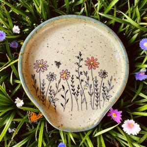 Op de afbeelding: Een keramische lepelhouder met een bloemenontwerp. De gebroken witte houder heeft een handgeschilderde illustratie van wilde bloemen in roze, geel en zwart, met een kleine bij. De rand is lichtblauw.