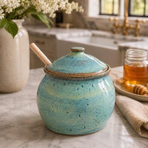 May include: A round, turquoise ceramic honey pot with a speckled finish and a matching lid. The pot features a brown rim and includes a light-coloured wooden honey dipper resting inside. The surface of the ceramic has a glossy, variegated blue-green glaze.