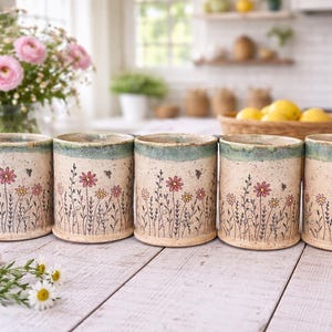 May include: Five ceramic mugs with a floral design. The mugs are cylindrical with a light beige base, a teal rim, and a handle on one. The floral design includes pink and yellow flowers, green stems, and small bees. The mugs are on a white wooden surface.