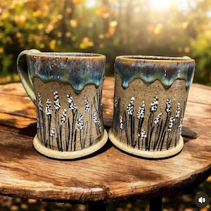 May include: Two ceramic mugs with a speckled tan base and a blue-green drip glaze at the rim. The exterior features hand-painted black stems and white floral details resembling lavender sprigs. Each mug has a cylindrical body and a flat base.