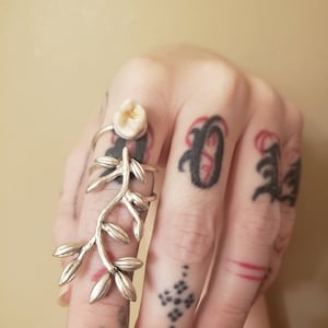 Puede incluir: Un anillo de plata con una muela en una banda, con un diseño separado en forma de vid que se extiende por el dedo. El anillo se lleva en una mano con varios tatuajes, incluyendo escritura negra y patrones geométricos.