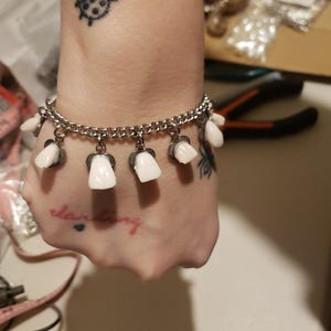 Puede incluir: Una pulsera de cadena plateada con dijes de dientes blancos. Un pequeño tatuaje de mariquita negra es visible en la muñeca. La palabra "darling" está tatuada en la mano.