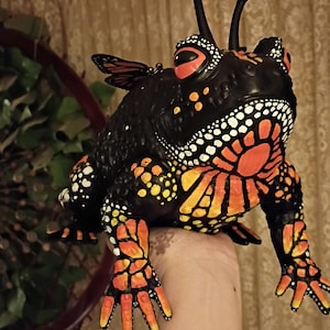Può includere: Figurina decorativa a forma di rana con dettagli di ali di farfalla. La rana è principalmente nera, con motivi arancioni e gialli sul corpo e sulle zampe. Ha macchie bianche, occhi rossi e due antenne nere.