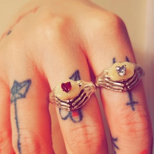 May include: Two silver rings with white stone settings. The rings are shaped like hands holding a heart. One ring has a red heart and the other has a clear heart.