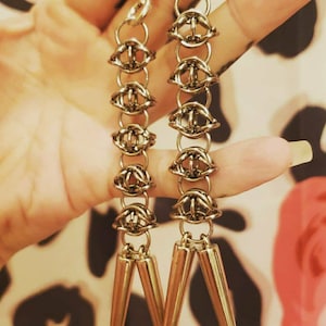 Puede incluir: Un par de pendientes de color dorado con un diseño de cadena y puntas puntiagudas. Los pendientes cuelgan de los lóbulos de las orejas.