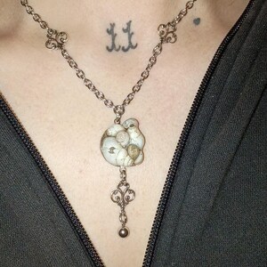 May include: A silver-toned necklace featuring a delicate chain with ornate filigree links. The pendant consists of a cluster of small, off-white spiral seashells arranged in a circular shape, with a dangling filigree charm and a small metal bead.
