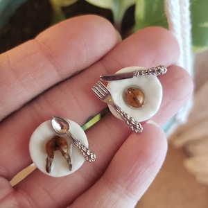 May include: Miniature white plates with a fried egg and a ladybug, and a pair of miniature plates with a spoon and fork. The plates have silver cutlery. The miniature food items are brown and tan.