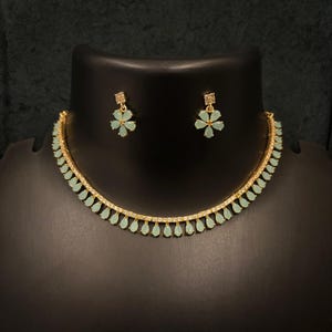 May include: A gold-coloured necklace and earring set. The necklace has small, light green teardrop-shaped stones and clear accent stones. The earrings feature a flower design with light green petals. The set is displayed on a black mannequin.