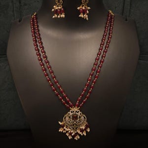 Puede incluir: Un conjunto de collar y pendientes. El collar presenta dos hilos de cuentas de color burdeos y un colgante dorado con una piedra roja central y detalles blancos. Los pendientes tienen un diseño similar, con cuentas burdeos y detalles blancos.