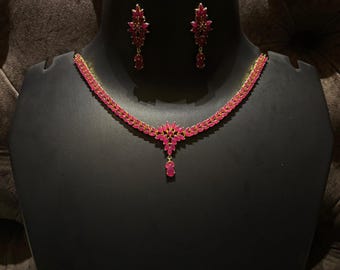 Puede incluir: Un conjunto de collar y pendientes de color rubí expuesto sobre un maniquí negro. El collar presenta un colgante floral y una cadena delicada. Los pendientes son de diseño floral a juego. La joyería se encuentra sobre un fondo oscuro texturizado.