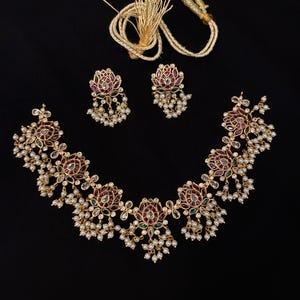 Puede incluir: Un conjunto de collar y pendientes en tono dorado con motivos florales y piedras rojas y verdes incrustadas. La joyería está decorada con pequeñas cuentas blancas tipo perla que cuelgan de los bordes. Incluye un cordón dorado con borla.