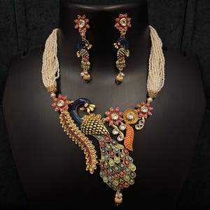 May include: A peacock-themed jewelry set featuring a necklace and matching earrings. The necklace has a central peacock pendant with blue, green, red, and gold accents, and a beaded strand. The earrings mirror the peacock design. The set is displayed on a black mannequin.