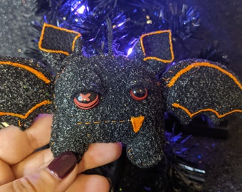 May include: A black glittery fabric bat ornament featuring orange stitched outlines on the wings and ears. The bat has one eye with a red and black circular design and a second eye with a red and black X pattern. A single orange tooth is visible.