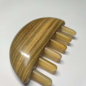 May include: A wooden hair comb with a half-moon shaped base and a row of beige teeth. The wood has a brown and green striped pattern.