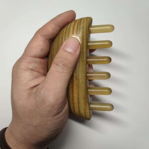May include: A wooden comb with a curved handle and five beige plastic teeth. The comb is held in a hand.