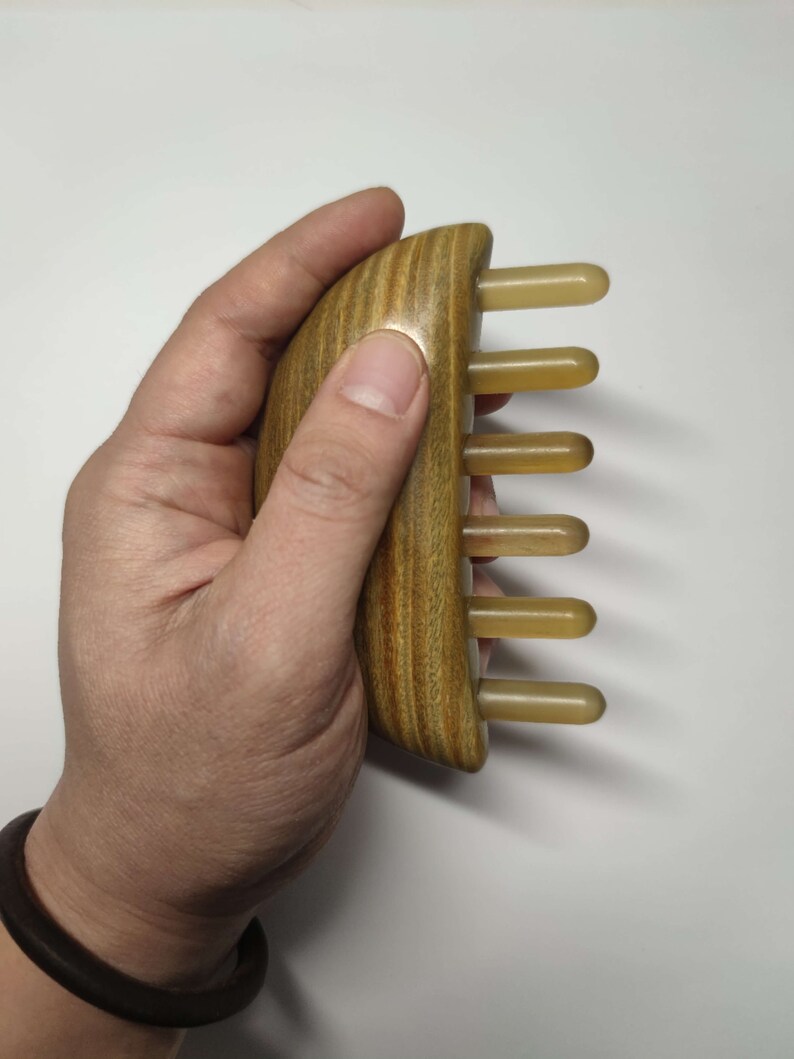 May include: A wooden comb with a curved handle and five beige plastic teeth. The comb is held in a hand.