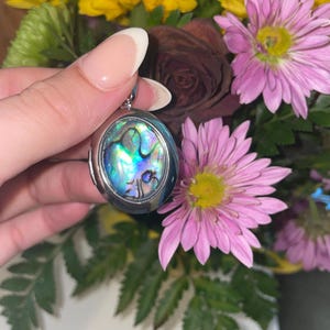 May include: Oval silver locket with iridescent abalone shell inlay, held in front of a bouquet of yellow and pink flowers. The locket's surface reflects shades of blue, green, and purple, creating a shimmering effect. The flowers include yellow chrysanthemums and pink daisies.