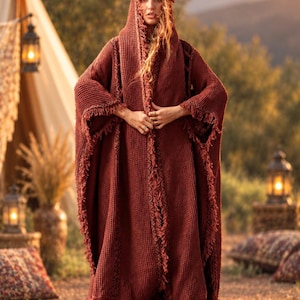 May include: A long, rust-colored hooded cloak featuring a textured waffle-knit fabric. The garment has an open front design with frayed, raw-edge detailing along the hood, sleeves, and vertical panels. The fabric has a consistent deep terracotta hue.
