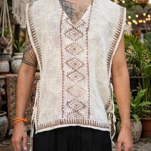 Peut inclure: Un gilet sans manches en tissu tissé texturé blanc cassé, doté d'une encolure en V et de broderies géométriques marron le long du centre et des bords. Le vêtement comporte des liens latéraux pour l'ajustement et une coupe droite ample.