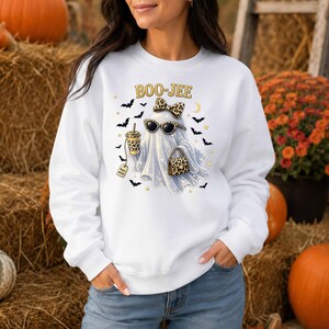 May include: A white long-sleeve sweatshirt featuring a graphic of a ghost wearing leopard print sunglasses and a matching bow. The ghost holds a leopard print coffee cup and handbag, surrounded by black bats, stars, and the text BOO-JEE.