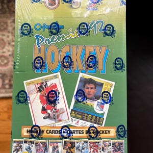 以下が含まれることがあります： 緑色の背景を持つ、未開封のO-Pee-Chee Premier 92ホッケーカードボックス。ボックスには「Premier 92 HOCKEY」、「HOCKEY CARDS CARTES DE HOCKEY」という文字が記載されており、2枚の大きなホッケーカードの画像と、小さなホッケー選手のカードが並んでいます。