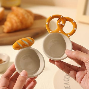Puede incluir: Dos cuencos de cerámica para mojar, uno con forma de pretzel y otro con forma de pan. El cuenco de pretzel es naranja con puntos blancos, y el cuenco de pan es marrón con detalles azules. Ambos cuencos tienen el interior color crema.