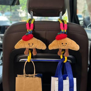 May include: Two plush croissant-shaped car seat organizers with smiling faces and red berets. They hang from the headrest with colorful cords and yellow hooks, holding shopping bags. The car seat is brown.