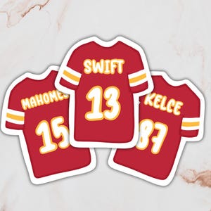 May include: Three red and white football jerseys with the names "Mahomes 15", "Swift 13", and "Kelce 87" printed on them.