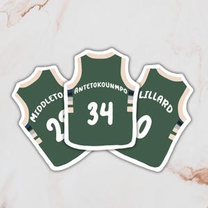 Puede incluir: Tres pegatinas de camiseta de baloncesto verde con números y nombres blancos. Las camisetas tienen rayas blancas y negras en los lados. Los nombres en las camisetas son "Middleton 22", "Antetokounmpo 34" y "Lillard 0".